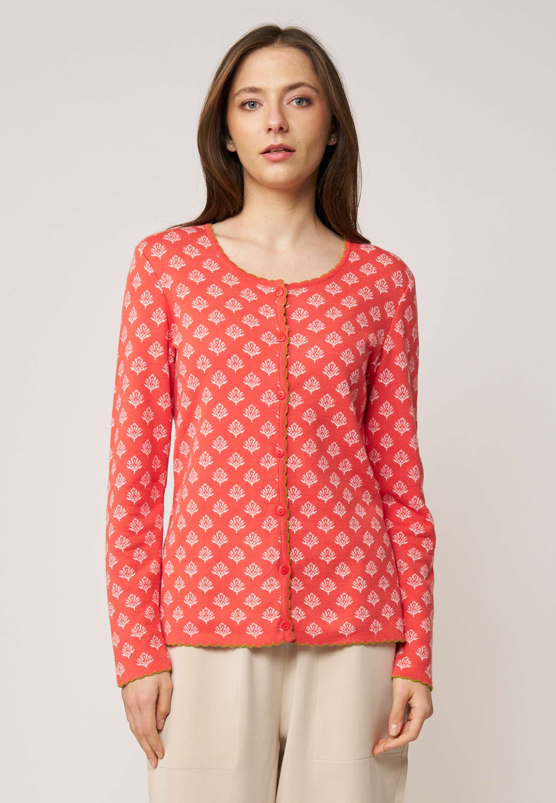 Frau trägt Strickjacke, Modell Lena, in Koralle mit weißem Muster und kontrastfarbener Kante vor hellem Hintergrund