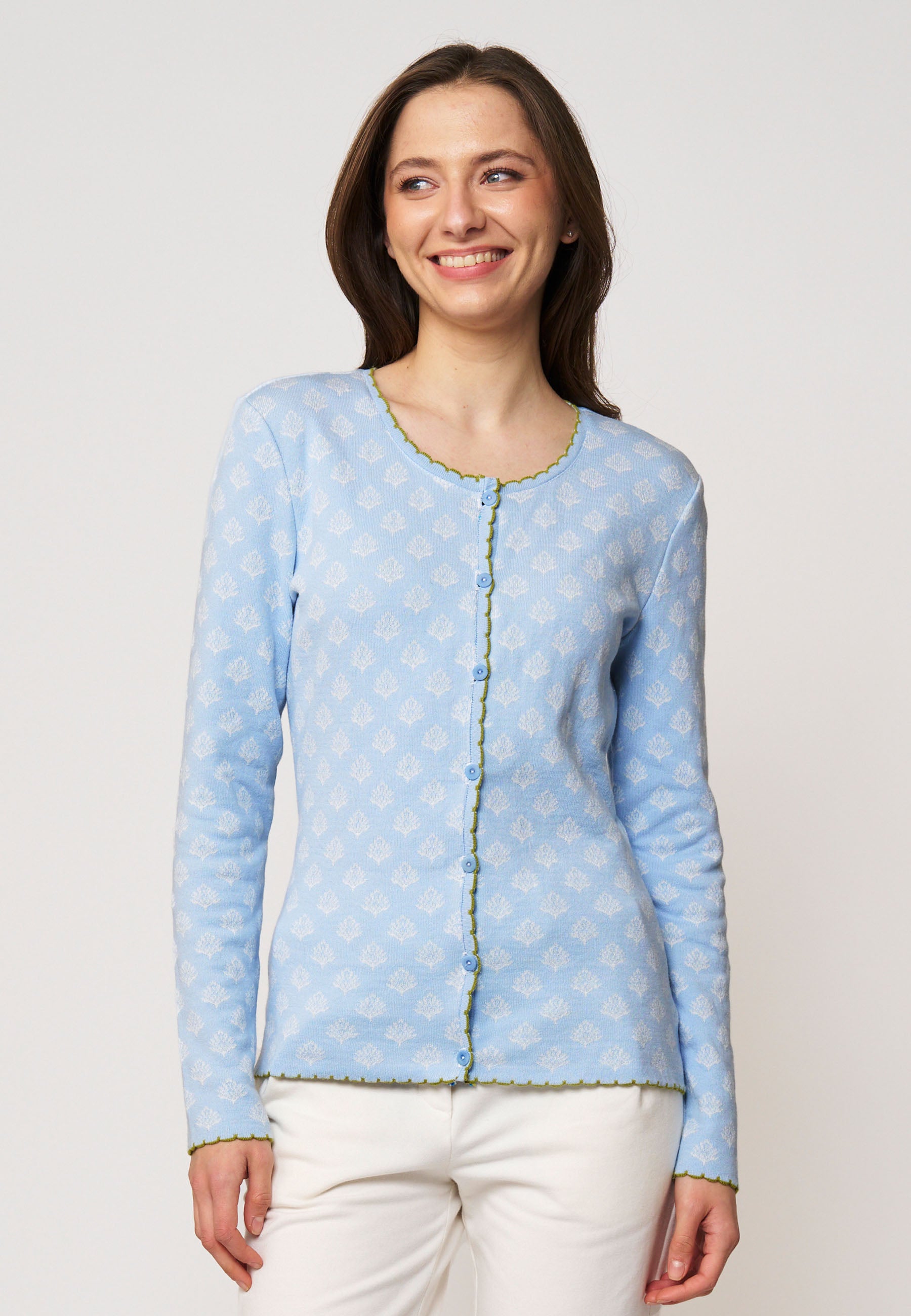 Frau trägt Strickjacke, Modell Lena, in hellblau mit zartem Muster und grünen Ziernähten, lächelnd vor weißem Hintergrund
